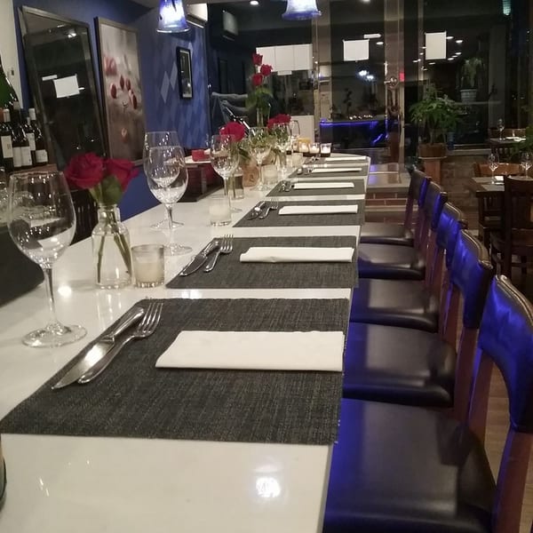A long dining table set with placemats, silverware, wine glasses, and red roses in a dimly lit restaurant.