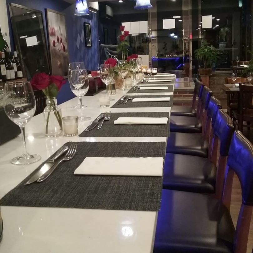 A long dining table set with placemats, silverware, wine glasses, and red roses in a dimly lit restaurant.