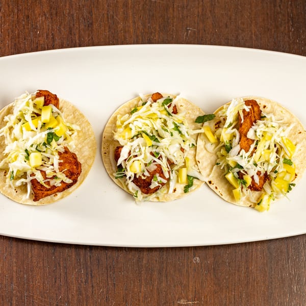 Three fish tacos with fried fish, shredded cabbage, and pineapple salsa on a white platter.
