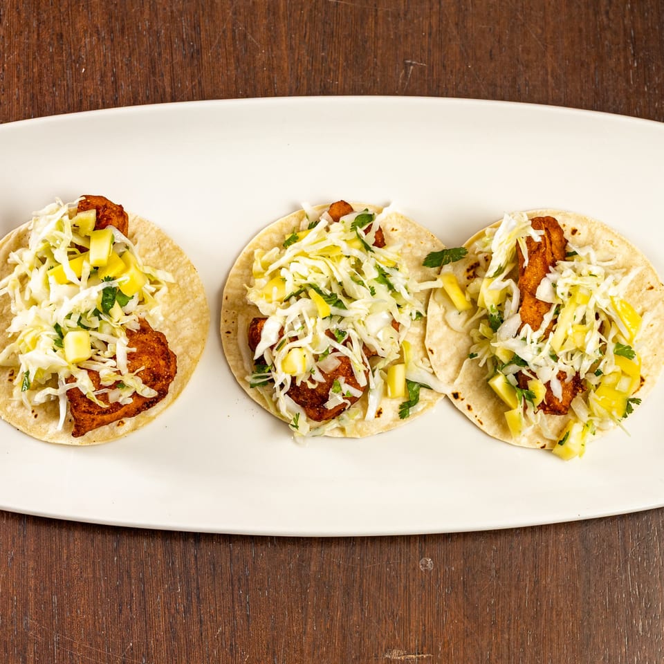 Three fish tacos with fried fish, shredded cabbage, and pineapple salsa on a white platter.