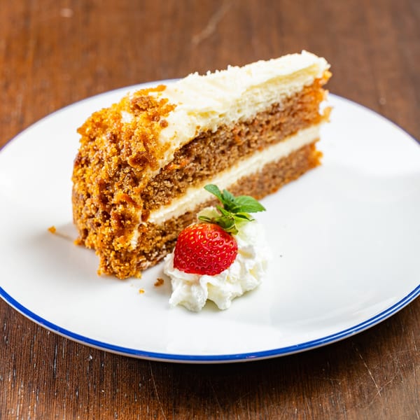 Slice of carrot cake with cream cheese frosting, topped with a strawberry, whipped cream, and mint.