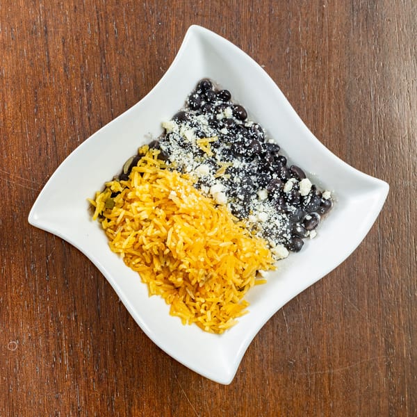 Mexican rice and black beans topped with crumbled cheese in a white bowl on a wooden table.