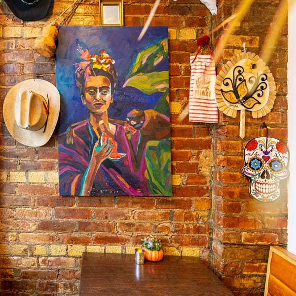 A vibrant Frida Kahlo painting and Mexican decor adorn a rustic brick wall above a wooden table.