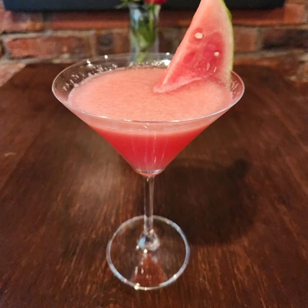 A pink watermelon margarita in a martini glass, garnished with a watermelon slice, on a wooden table.