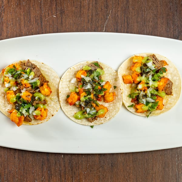 Three Bistro Casa Azul tacos with meat, sweet potato, cilantro, and green sauce on a white plate.