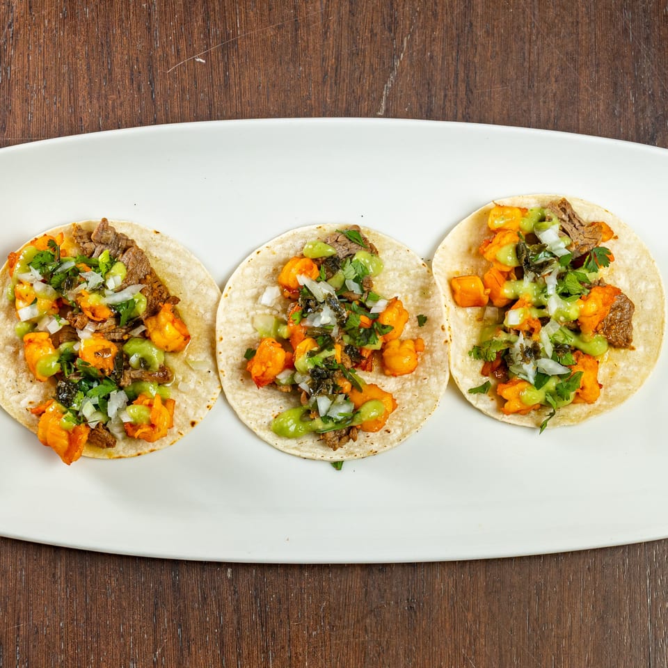 Three Bistro Casa Azul tacos with meat, sweet potato, cilantro, and green sauce on a white plate.